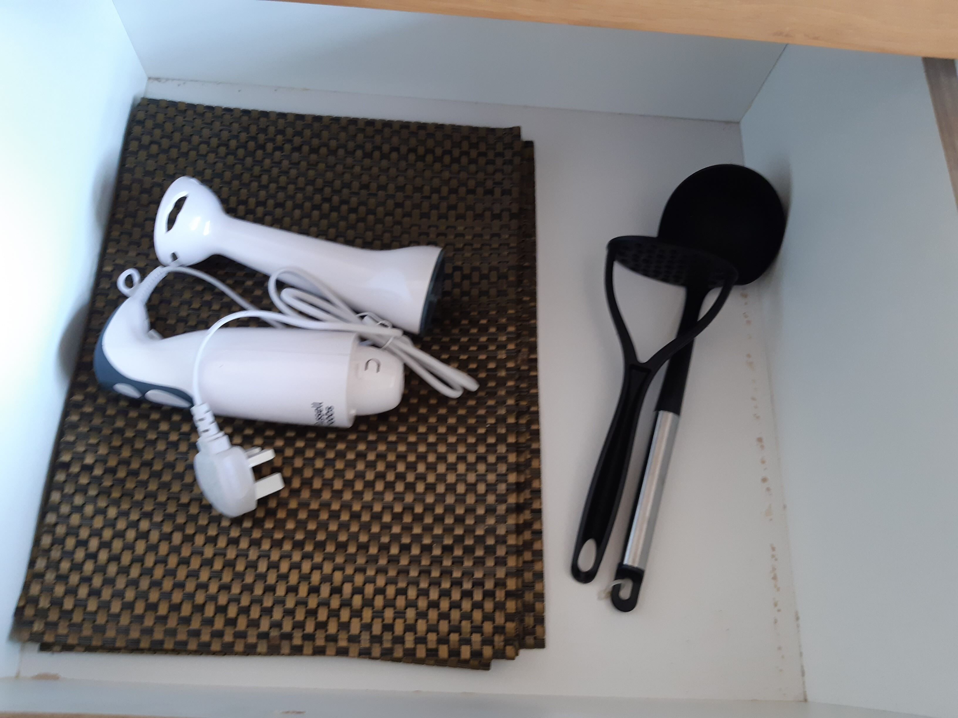 A white box containing a white hairdryer, a black and silver spatula, and a white power adapter on a woven mat.