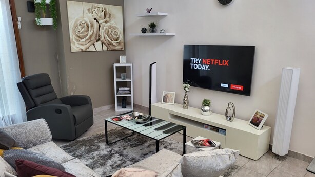 A modern living room with a grey sofa, a black recliner, a coffee table, a TV displaying a Netflix advertisement, a white console table, and a wall-mounted clock. - Accra Cozy Retreat With 2 Bedrooms (Santeo)