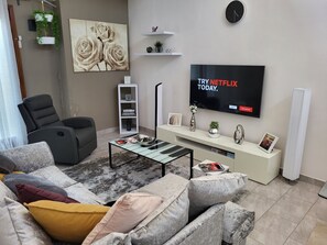 A modern living room with a grey sofa, a black recliner, a coffee table, a TV displaying a Netflix advertisement, a white console table, and a wall-mounted clock. - Accra Cozy Retreat With 2 Bedrooms (Santeo)