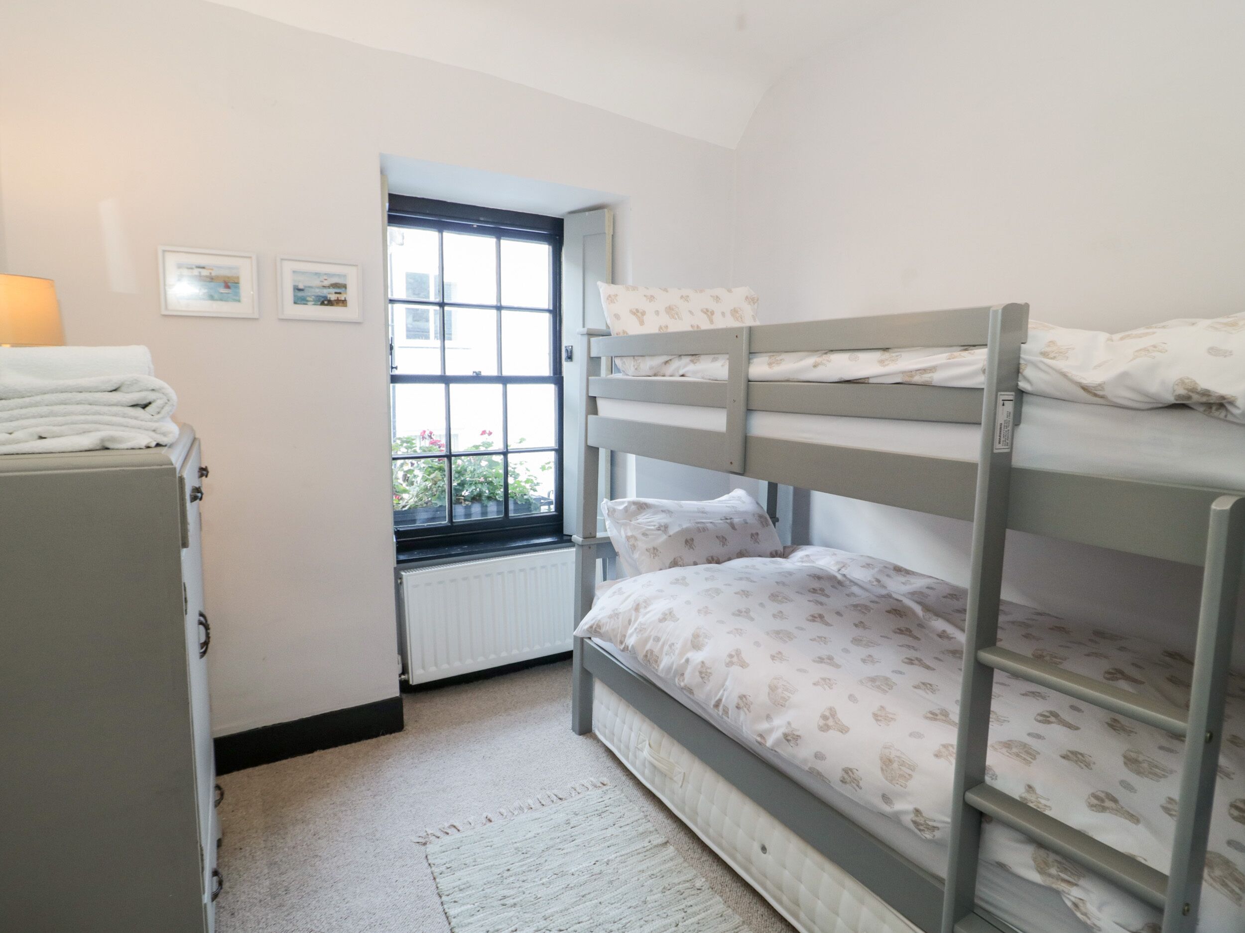 A bunk bed room with a radiator, a window with a view of greenery, and a small desk with a lamp.