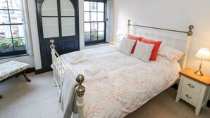 A neatly arranged bedroom with a bed, bedside tables, a bench, and a window with a view of buildings and flowers. - Sea Chest (Conwy)