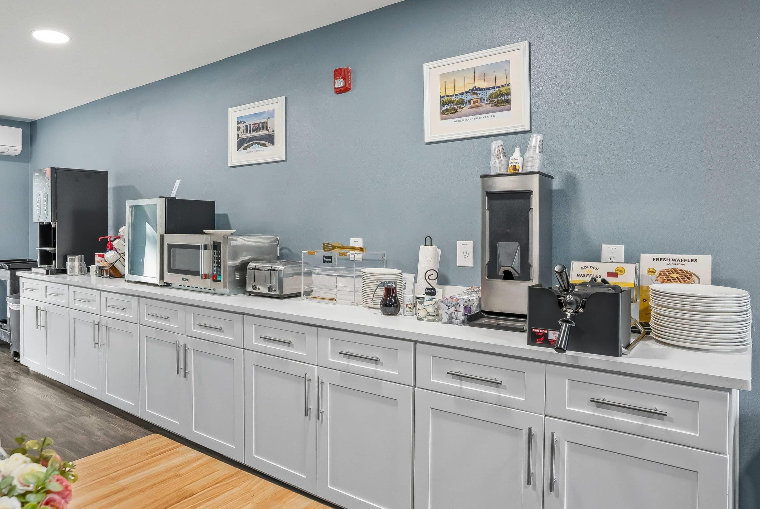 A modern kitchen area with white cabinets, a microwave, toaster, and waffle maker. There are framed pictures on the wall and a fire extinguisher mounted above the counter.