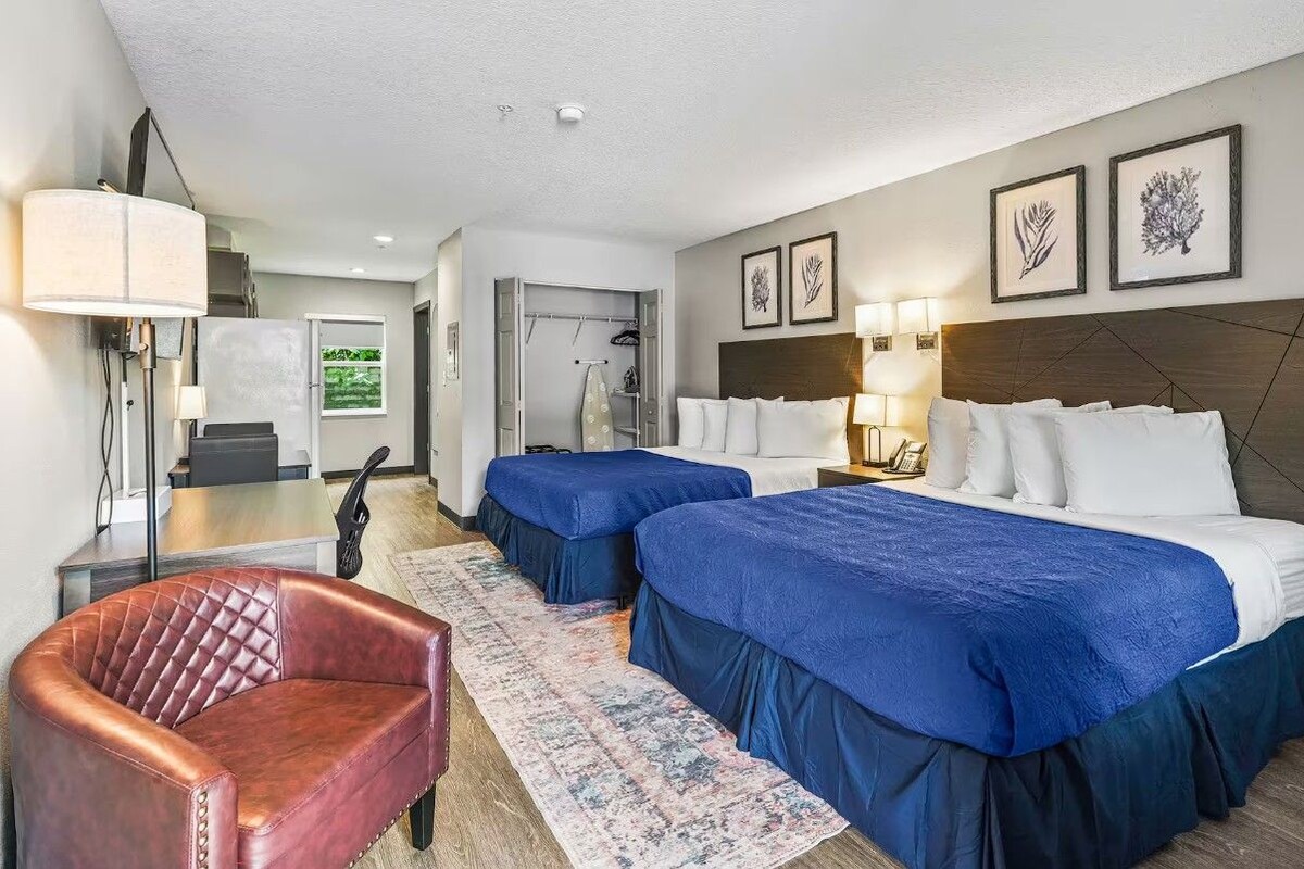 A hotel room with two beds, a red leather chair, a desk, and a large window.
