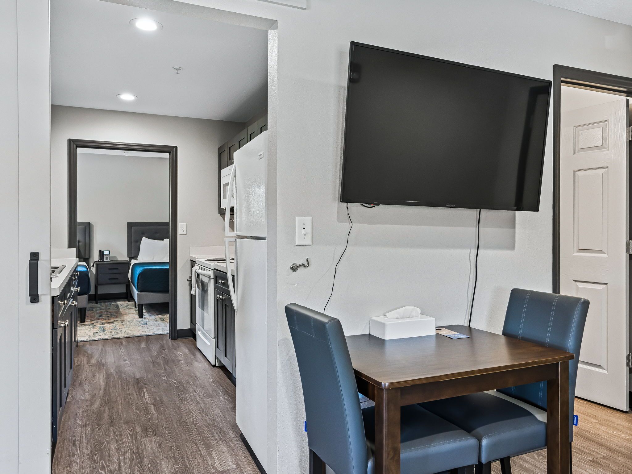 Habitación de hotel compacta con kitchenette, zona de comedor con dos sillas, televisor de pantalla plana montado en la pared y una puerta que da a otra habitación.