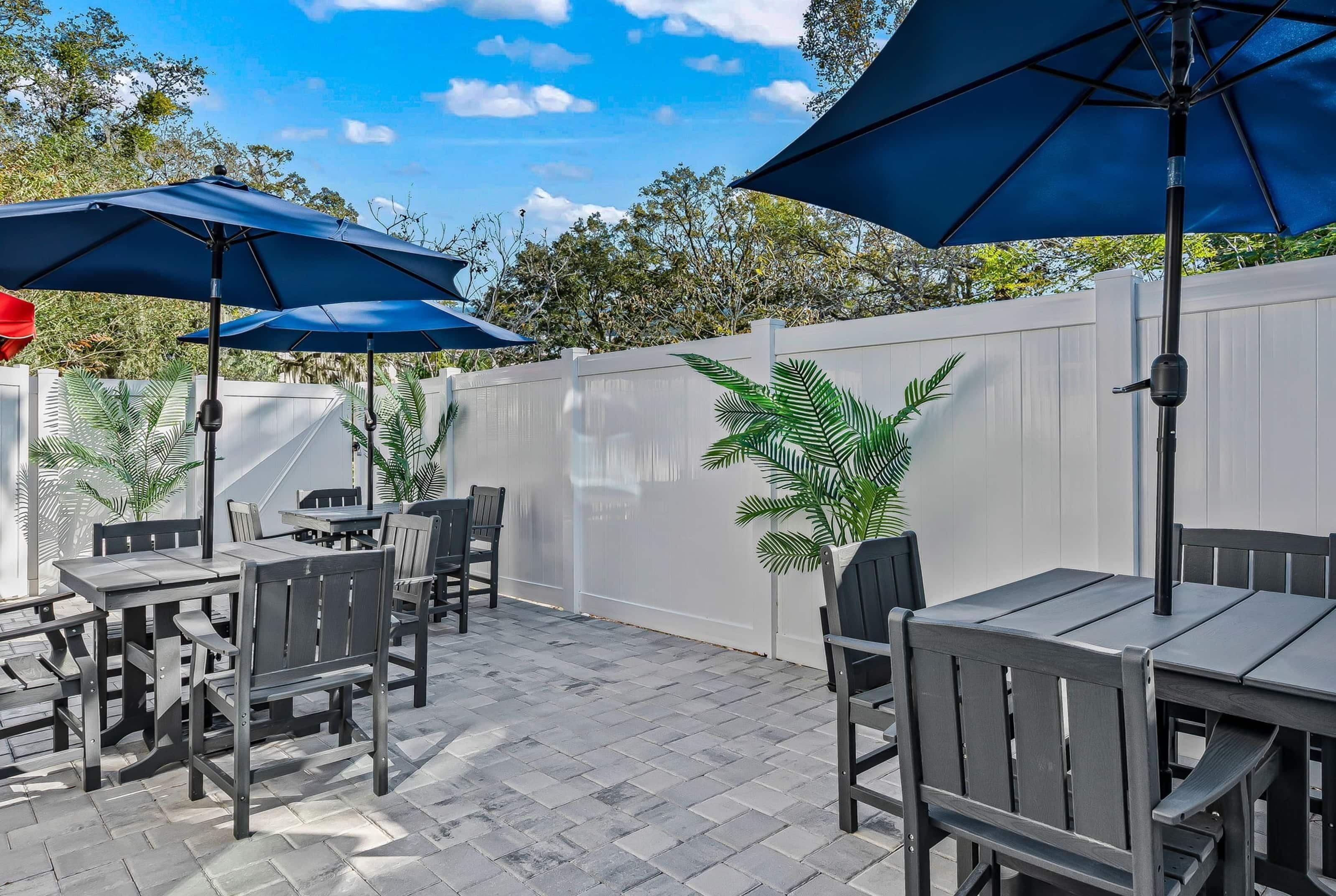 An outdoor patio with tables and chairs, blue umbrellas, and a white fence.