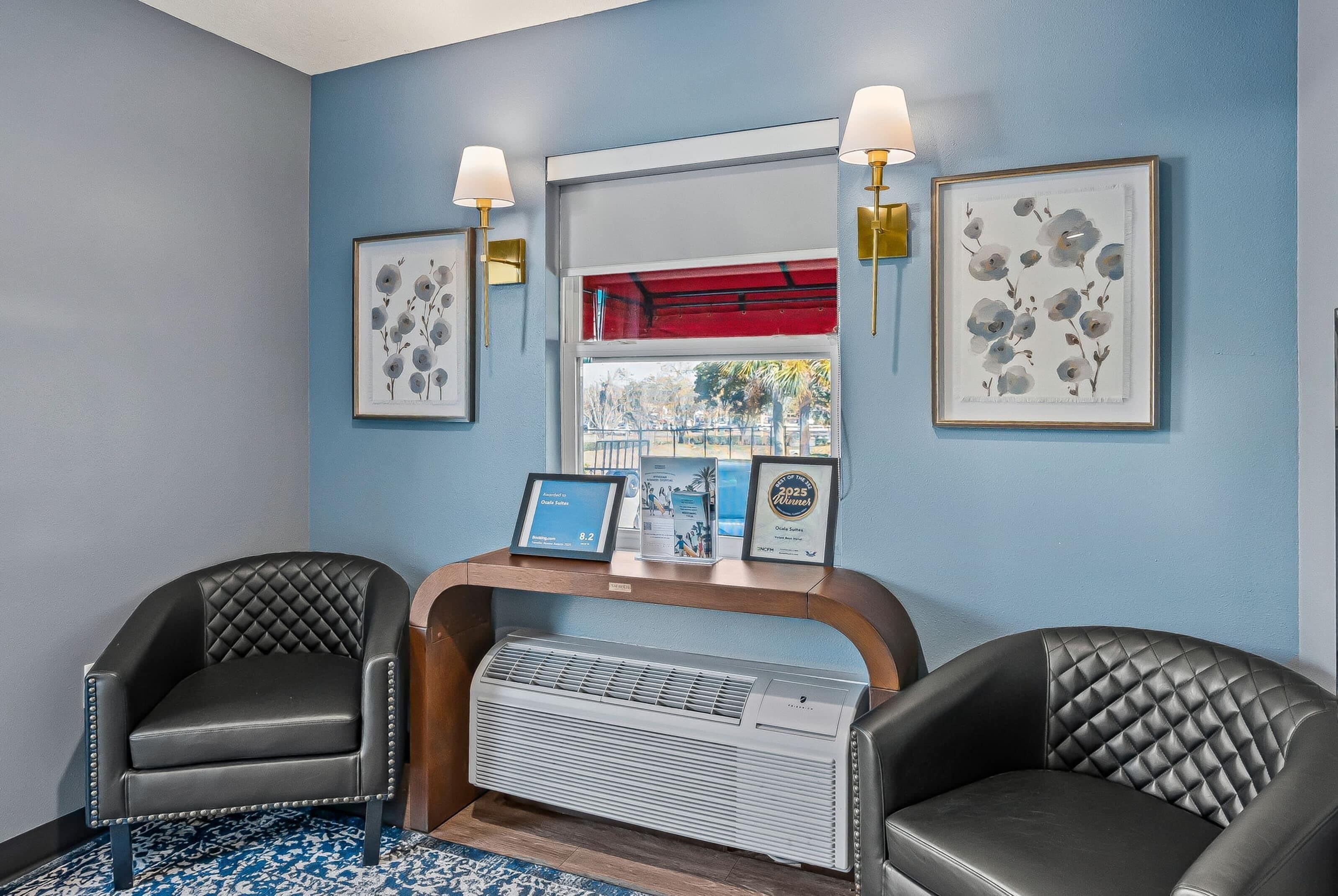 A room with two black leather chairs, a wooden console table with framed artwork, a large air conditioning unit, and a window with a red blind.
