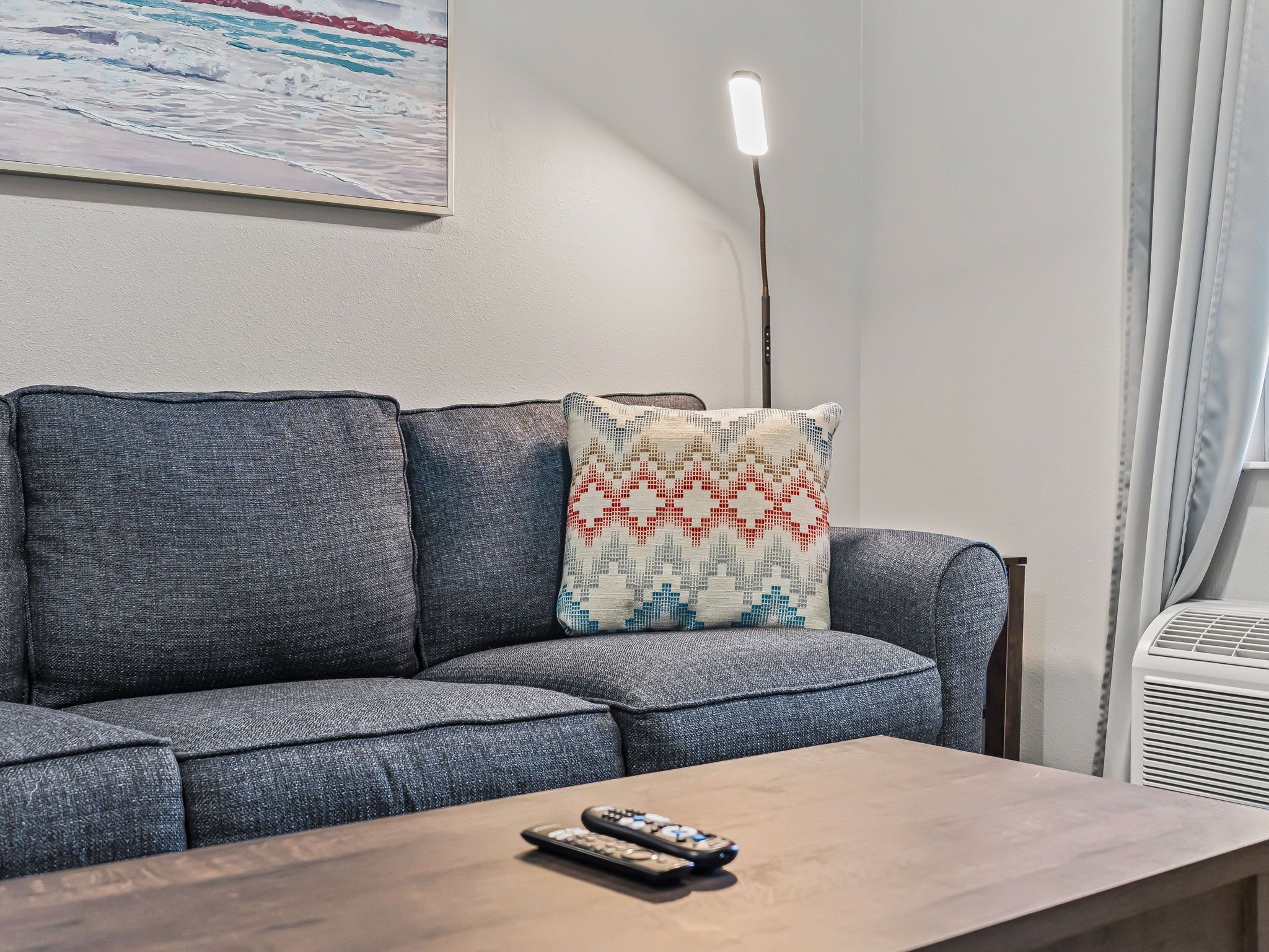 A grey sofa with a patterned throw pillow, a wooden coffee table with a remote control, and a painting of a seascape on the wall.
