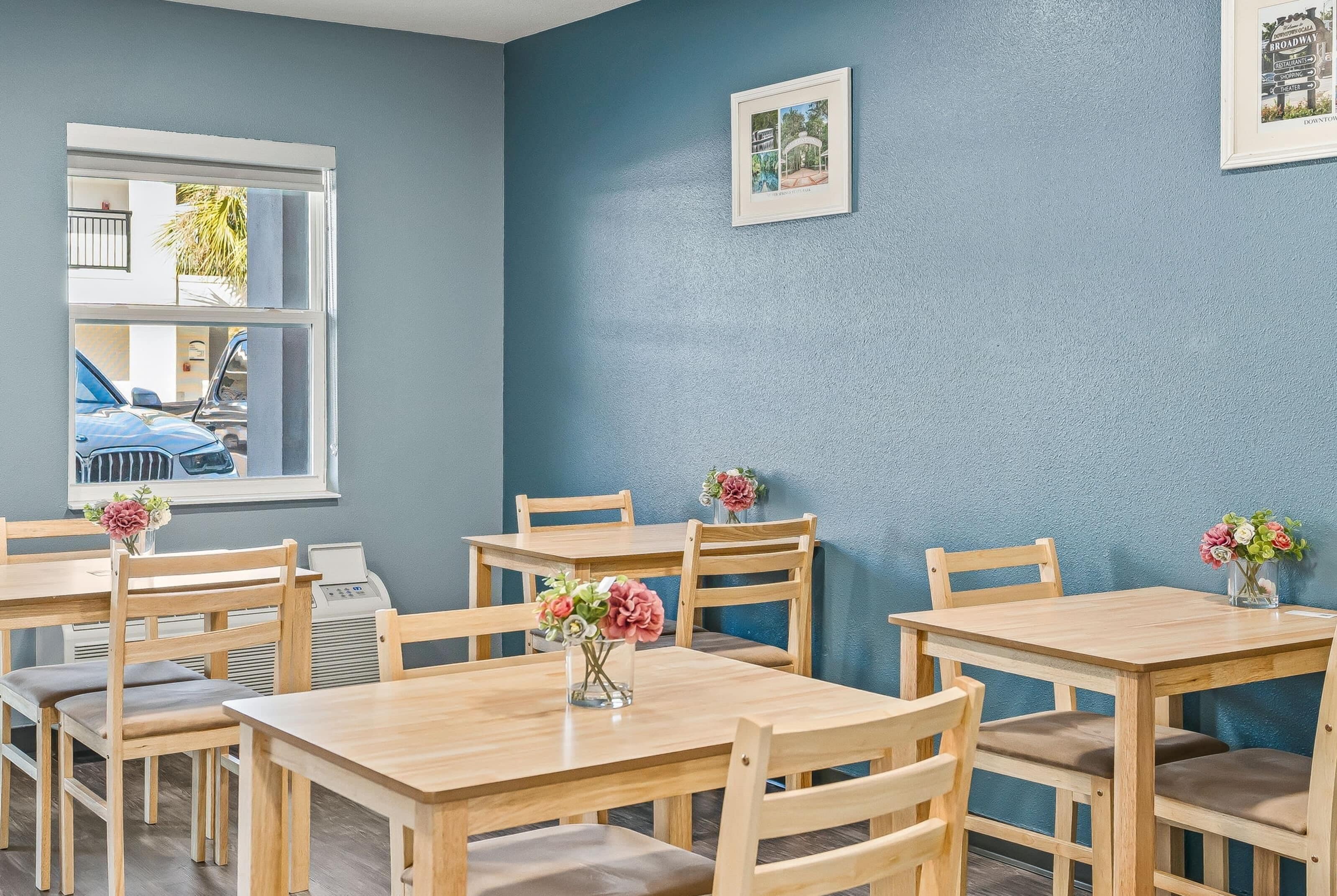 A dining area with wooden tables and chairs, a window with a view of parked cars, and a framed picture on the wall.