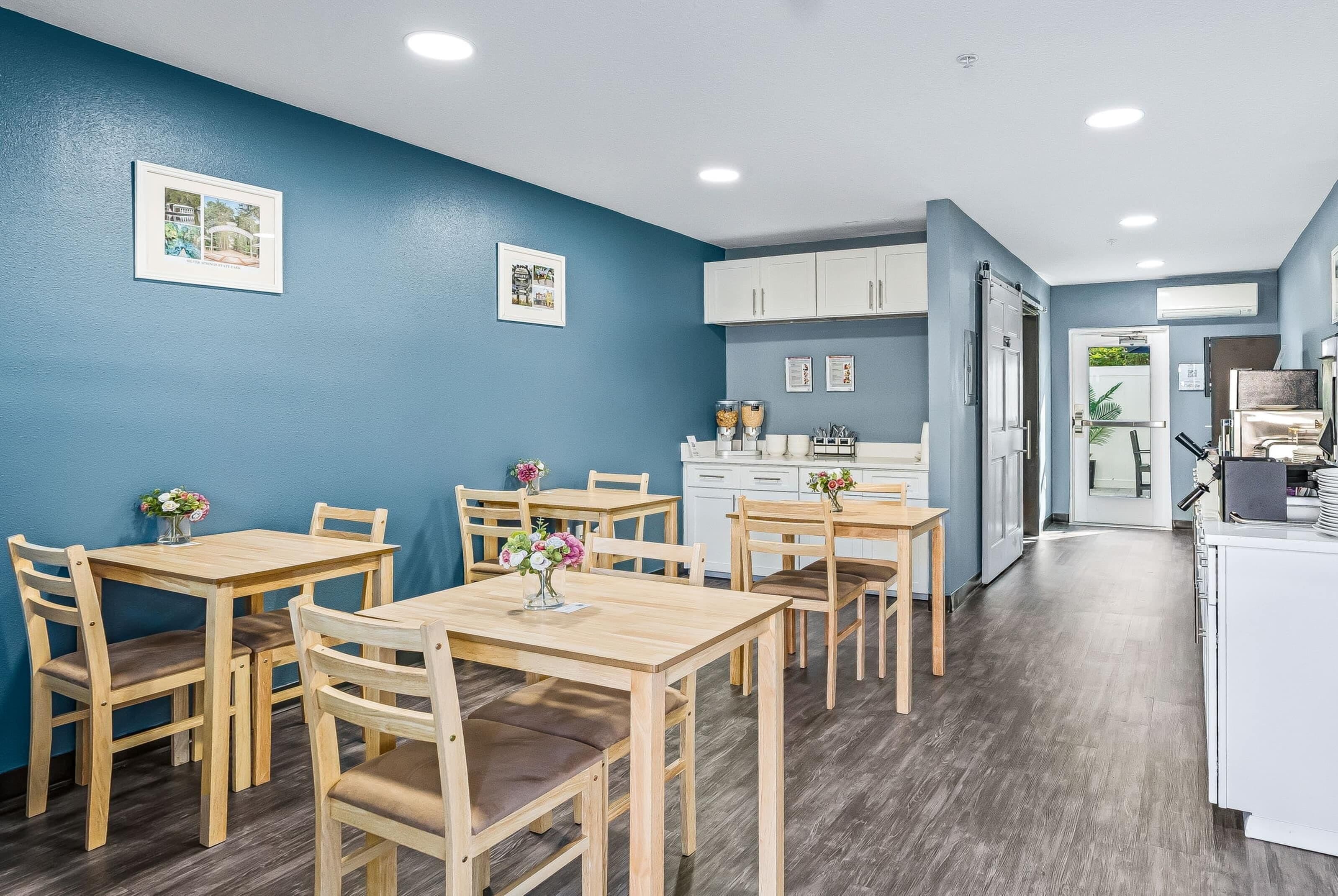 Un comedor con mesas y sillas de madera, una zona de cocina con armarios blancos y un pasillo que lleva a una puerta.