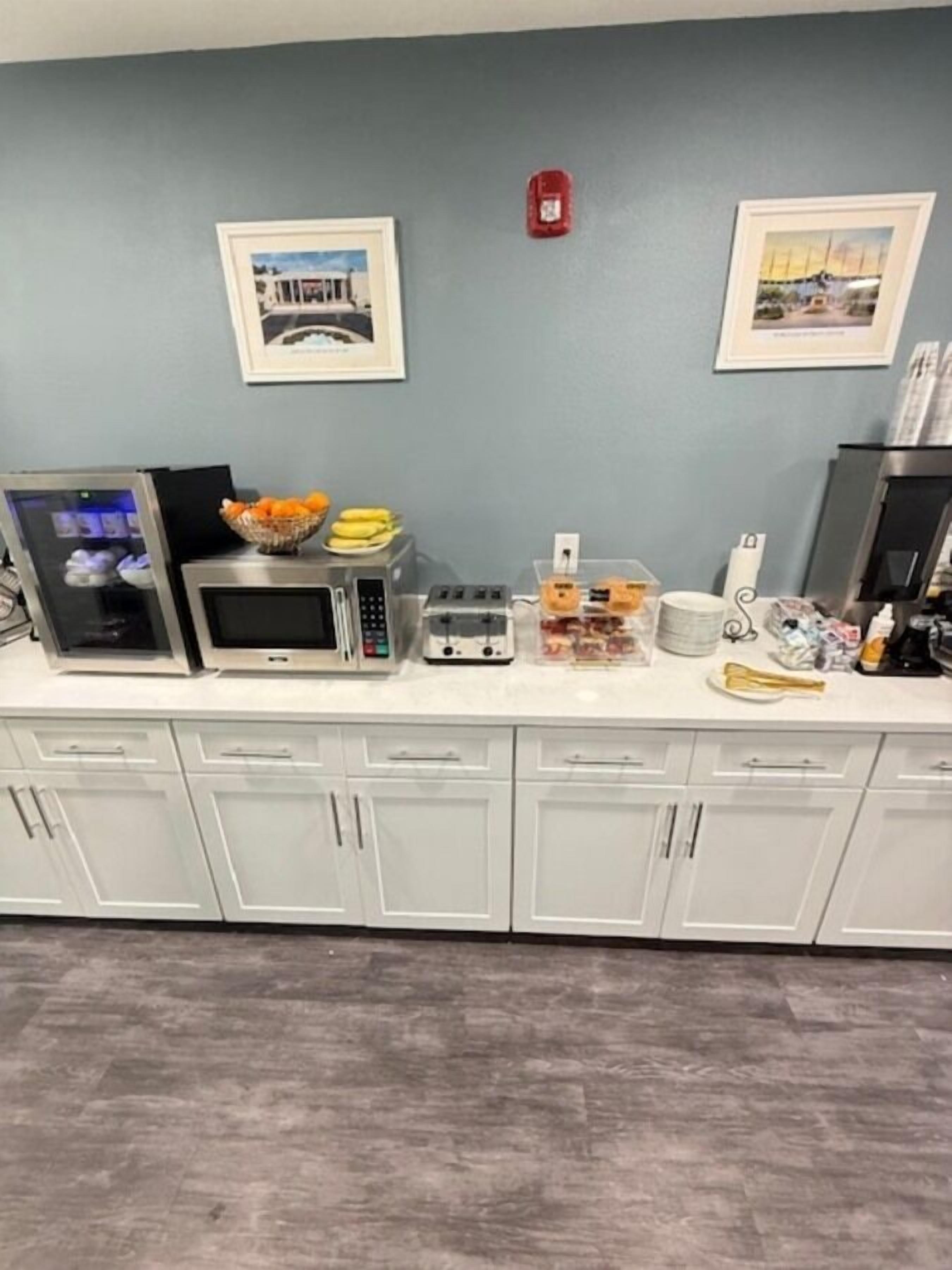A kitchen with white cabinets, a microwave, toaster, and various food items on the counter. There are two framed pictures on the wall and a fire alarm.