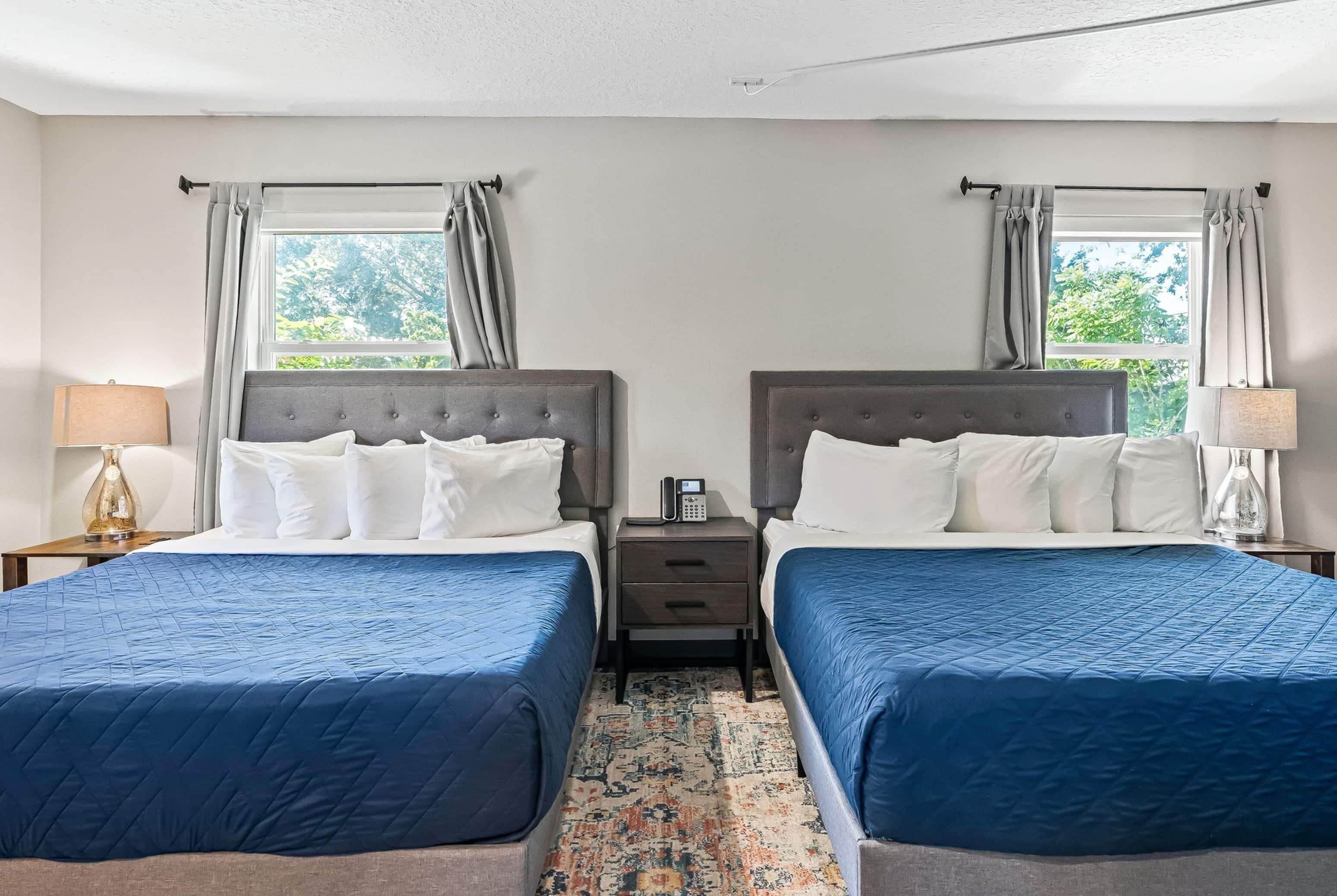 Habitación doble con dos camas, cada una con cubrecama azul acolchado, una mesita de noche pequeña con lámpara y teléfono.