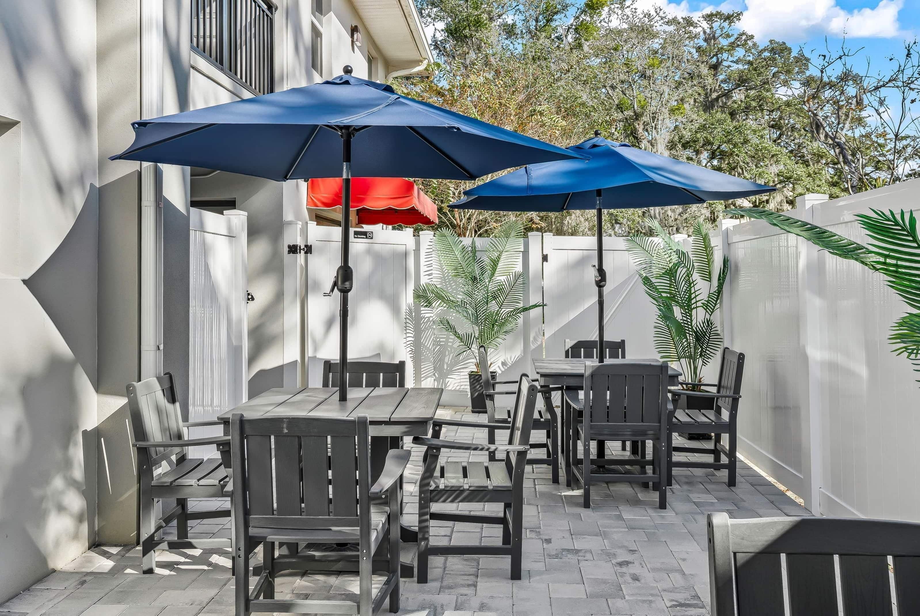 Une terrasse extérieure avec des tables et des chaises en bois, deux parasols bleus et des plantes en pot.