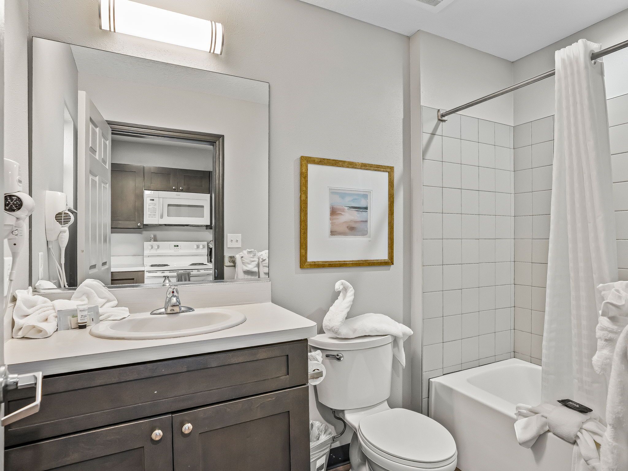 Un baño moderno con lavamanos, tocador y retrete blancos. Se aprecia una cortina de ducha, un cuadro enmarcado en la pared y un perchero para toallas.