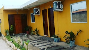 A yellow building with a covered entrance, potted plants, and a paved walkway. - Chalés Pôr do Sol Gostoso (São Miguel do Gostoso)
