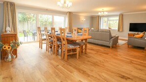 A spacious living area with a wooden dining table, chairs, and a grey sofa. There is a TV and a window with curtains. - Fallowfield (Great Yarmouth)