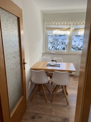 A dining area with a wooden table and white chairs, a window with frosted glass, and a view of snow-covered trees.