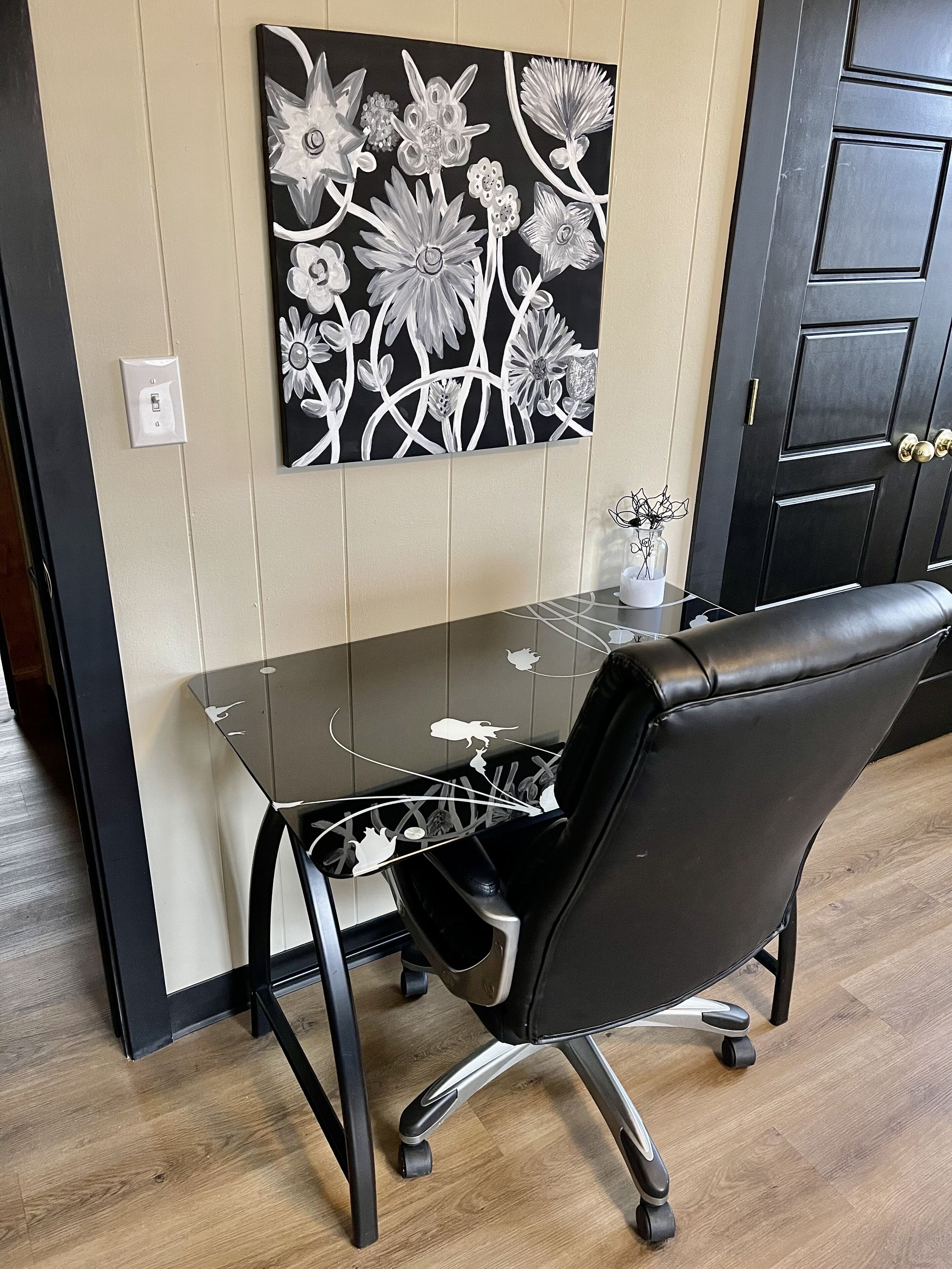 A modern office space with a glass desk, black leather office chair, and a wall-mounted floral artwork.