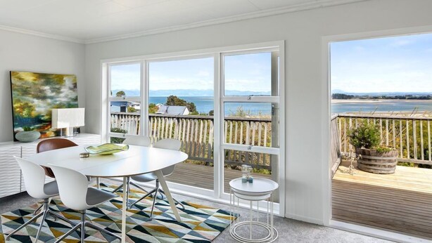 A bright room with a large window overlooking a beach and ocean. There is a white table with chairs, a small round table, and a colorful rug. - Emily's Beach House - Kaiteriteri Holiday Home (Kaiteriteri)