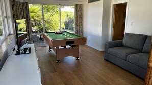 A room with a pool table, a grey sofa, and a window with a view of greenery. - Emily's Beach House - Kaiteriteri Holiday Home (Kaiteriteri)