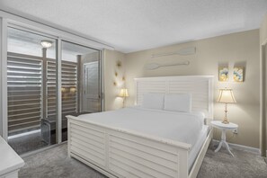 A bedroom with a large bed, a sliding glass door to a balcony, a white headboard, and two bedside lamps.