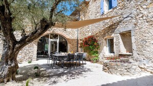Un espace extérieur aménagé pour les repas, avec des murs en pierre, un grand arbre et une table entourée de chaises.