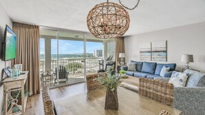 Un soggiorno spazioso con una grande finestra che offre vista sulla spiaggia e sullo skyline della città. Si trova un divano confortevole, un tavolo da pranzo e un tavolino da caffè in vimini.