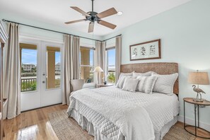 Une chambre à coucher avec un grand lit, un ventilateur de plafond, une fenêtre avec des rideaux et un tableau encadré au mur.