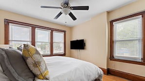 A bedroom with a bed, a ceiling fan, a flat-screen TV, and large windows with blinds.