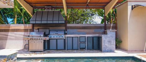 An outdoor kitchen with a grill, sink, and storage cabinets, adjacent to a swimming pool.
