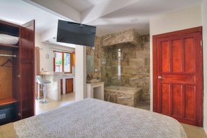 Quarto com cama, televisão fixada na parede, banheiro com chuveiro e porta de madeira.