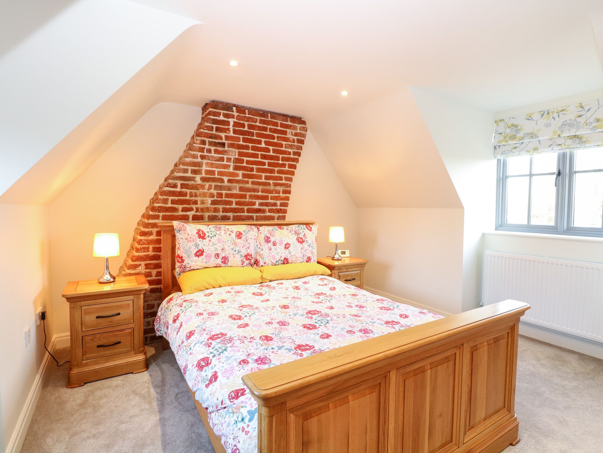 A bedroom with a sloped ceiling, a brick wall, a wooden bed, two bedside tables with lamps, and a window with a floral curtain.