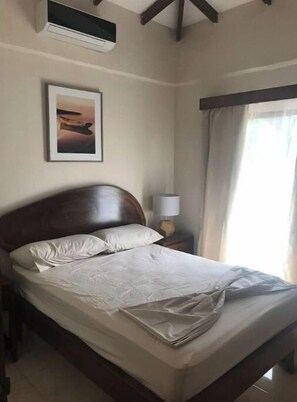 A neatly made bed in a hotel room with a headboard, a framed picture on the wall, a lamp on a bedside table, and a window with curtains.