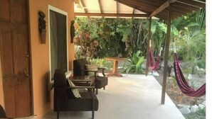 Un espacio al aire libre techado con sofá y sillas, una puerta y vistas a un jardín con una hamaca.