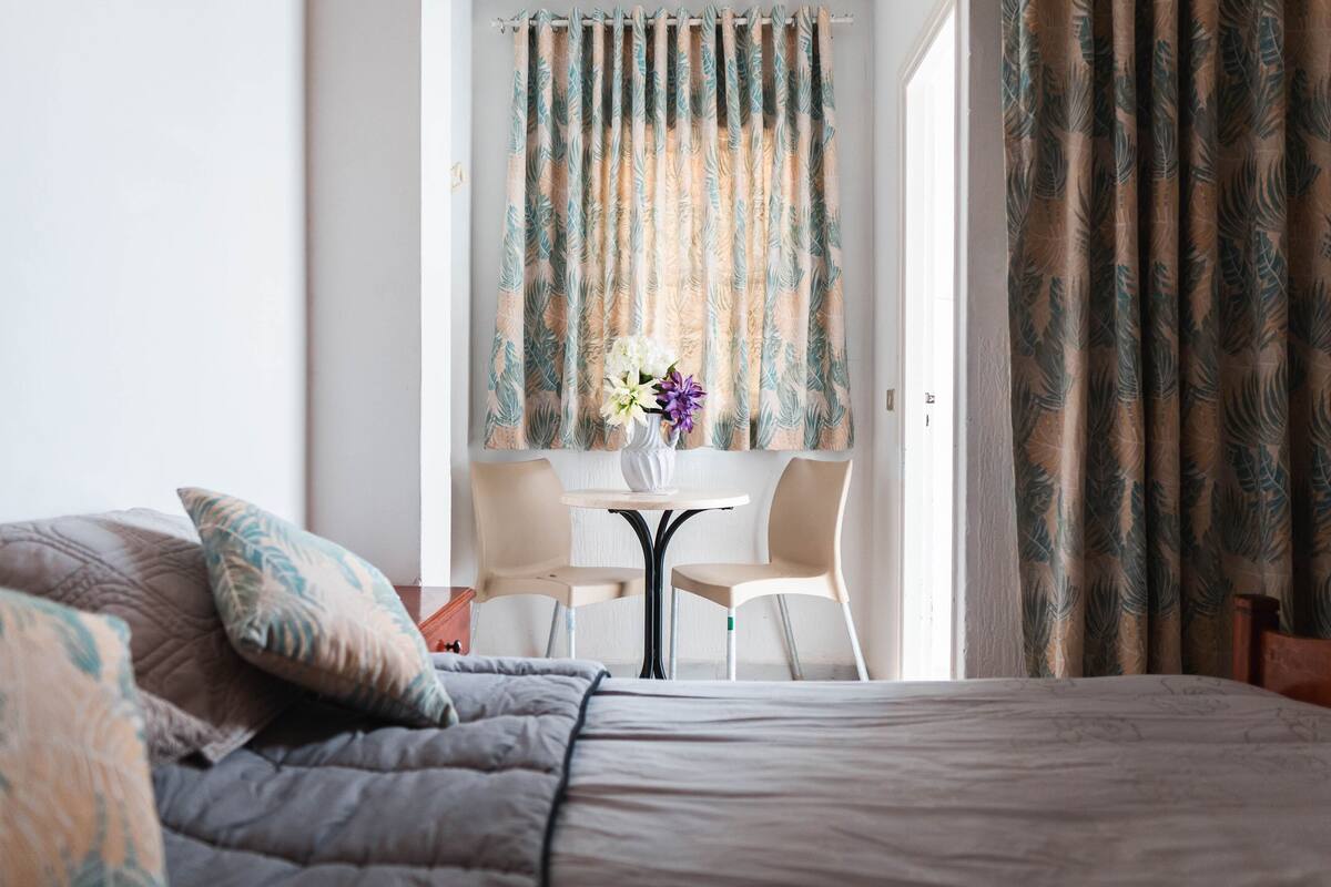 A bedroom with a bed, pillows, a small round table with chairs, and curtains.