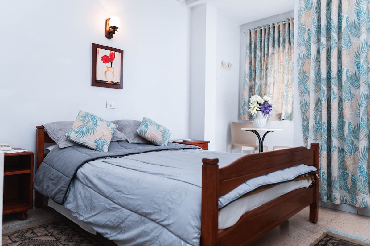 A bedroom with a bed, bedside tables, a nightstand, a small table with flowers, and a window with curtains.