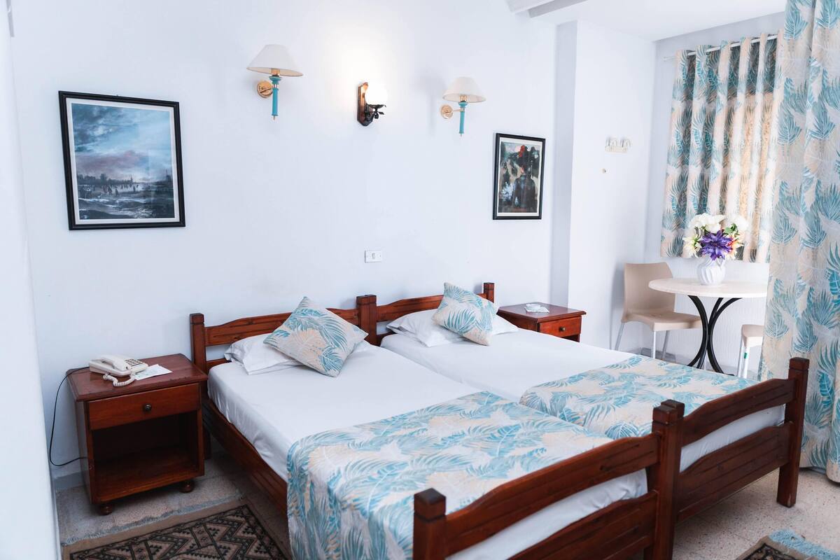 A double bed with a leaf-patterned bedspread, a bedside table with a phone, and a wall-mounted lamp.