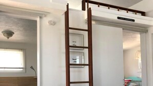 A room with a bed, a white round table with four white chairs, a wooden ladder, and a window with blinds. - Quarterboard 9 (Sandwich)