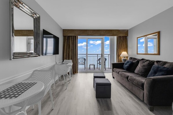 Een moderne woonkamer met een grijze bank, een witte tafel en stoelen, een balkon met uitzicht, en een grote spiegel aan de muur.