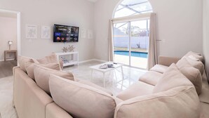 A spacious living room with a large TV, a white sofa, a coffee table, and a view of a swimming pool through a glass door. - Cozy on Cavendish Themed Bedrooms (Kissimmee)