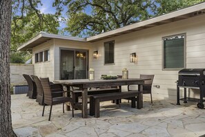 A patio with a stone floor, a wooden table with chairs, a barbecue grill, and a house with a white exterior and large windows.