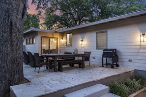 An outdoor patio with a wooden table and chairs, a barbecue grill, and a stone-tiled floor.