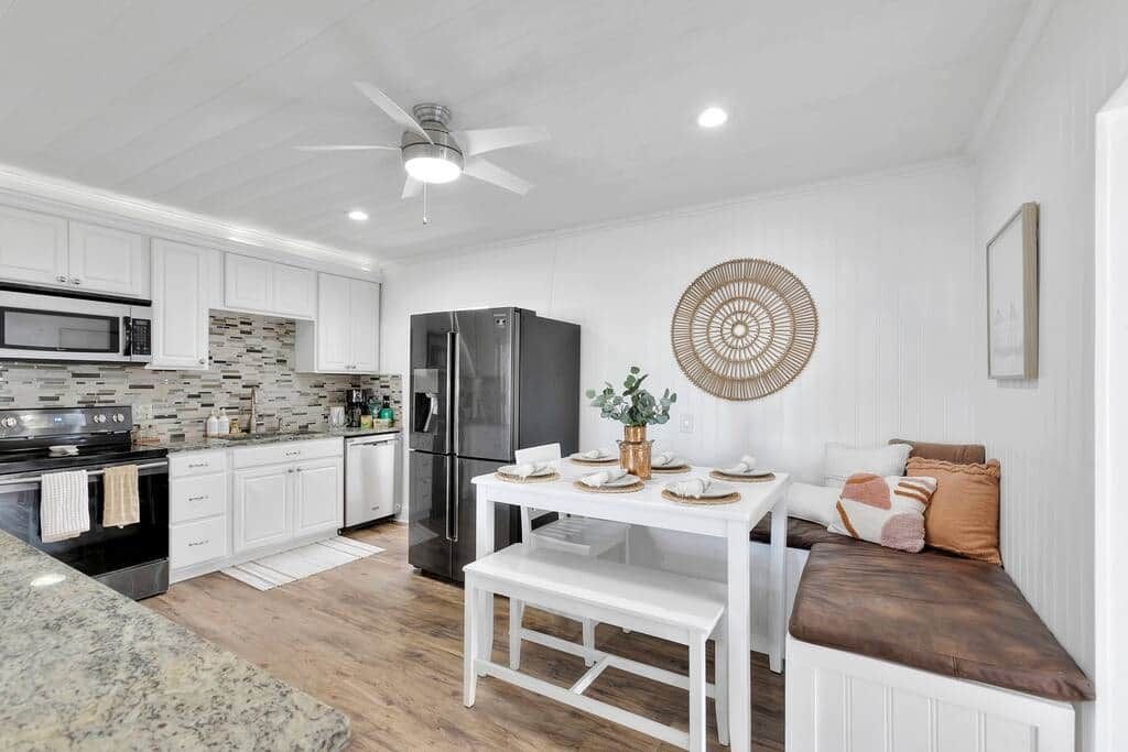 Una cocina moderna con armarios blancos, un refrigerador negro y un comedor con una mesa y un banco blancos.