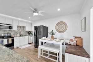 A modern kitchen with white cabinets, a black refrigerator, and a dining area with a white table and bench. - Bungalow! Hot TUB! Games! 10 min to Beach! (Palm Beach County)