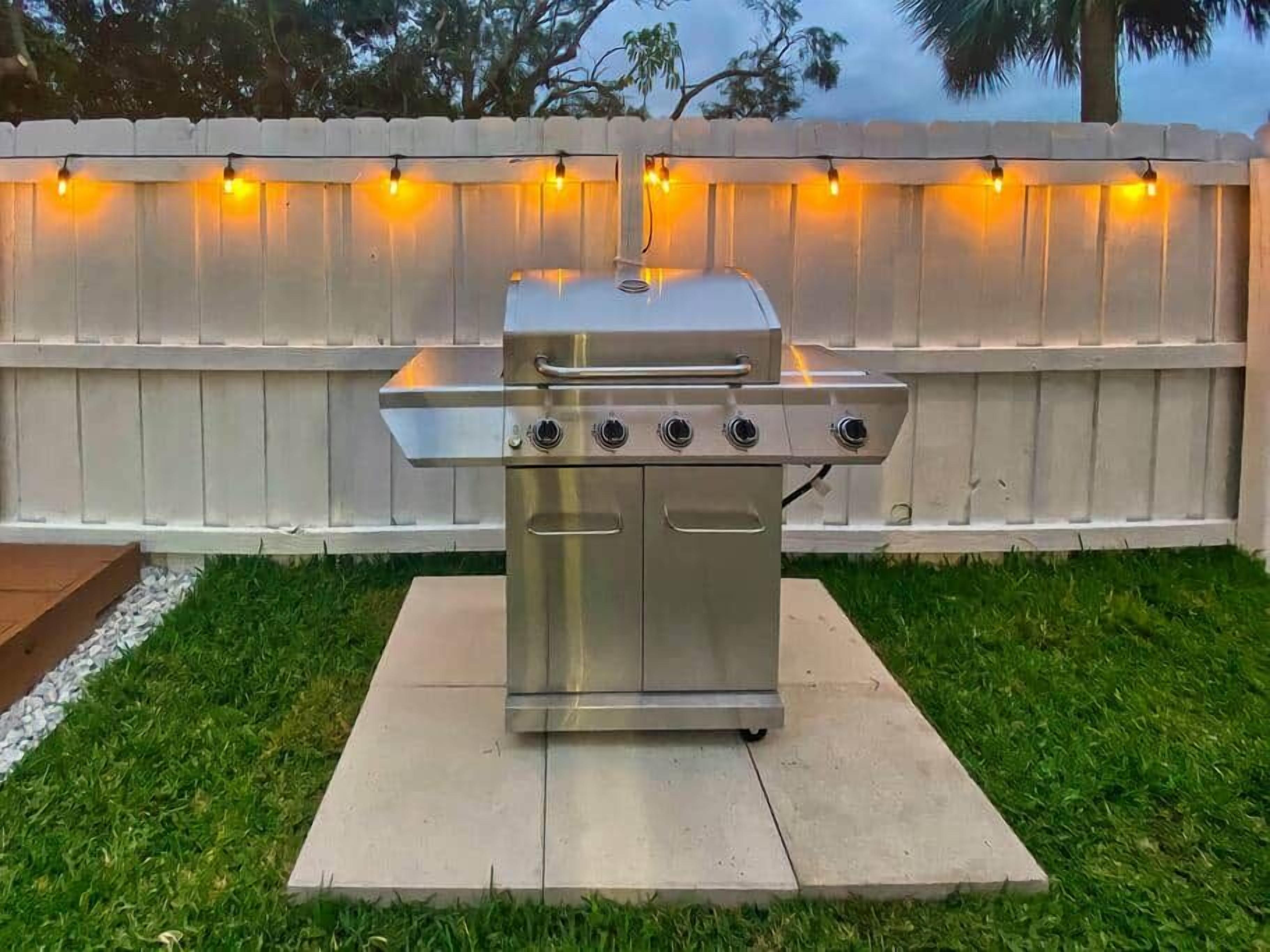 A stainless steel barbecue grill with four burners, a handle, and a lid, placed on a concrete pad in a grassy backyard.