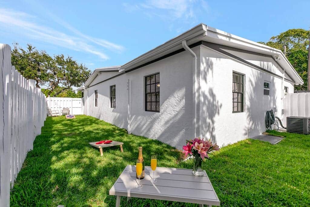 A white house with a fenced backyard, a table with drinks and flowers, and a clear sky.