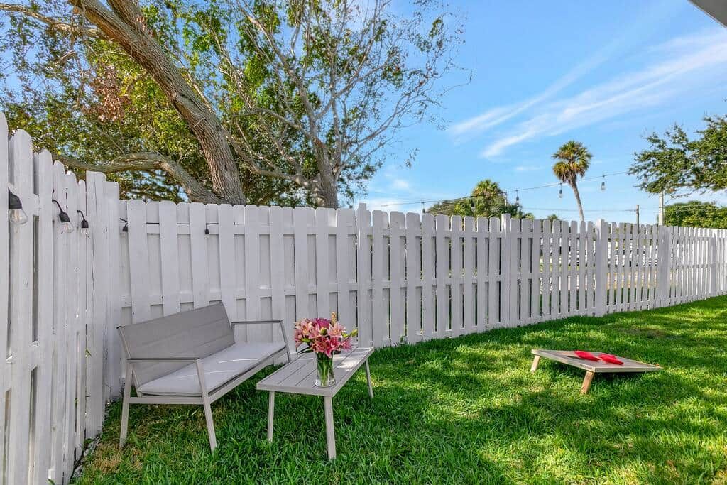 A white picket fence, a bench, a table with a vase of flowers, and a small table with a red object.