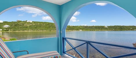 A room with two large arched windows overlooking a lake, a chair with a striped cushion, and a view of a hillside with buildings.