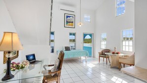 Una habitación luminosa y amplia con cama, escritorio con computadora portátil, silla, mesa con un florero y ventanas grandes con vistas al mar.