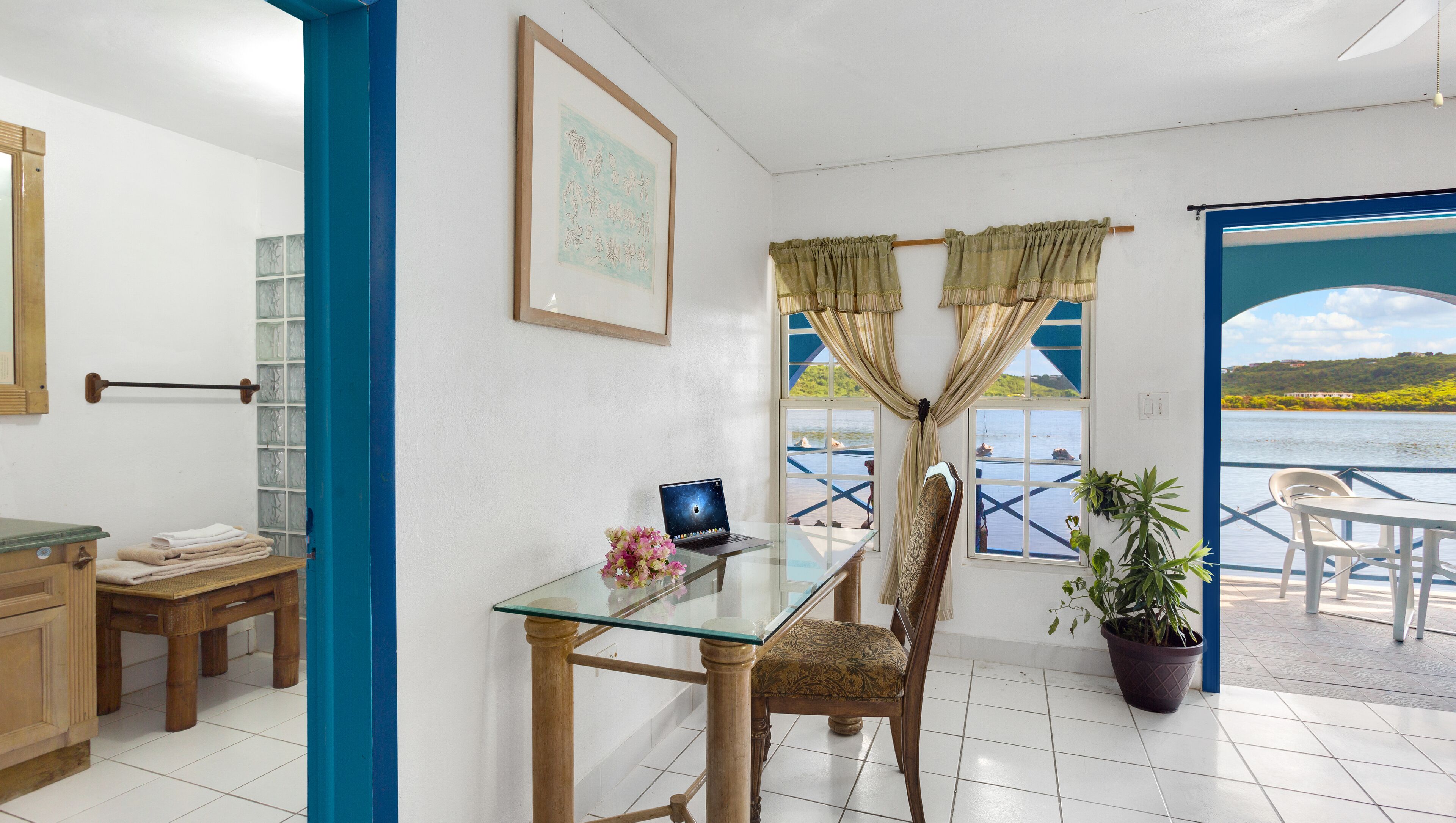 Una habitación con una mesa de vidrio, una computadora portátil, una silla, una planta en maceta y vistas a un cuerpo de agua.