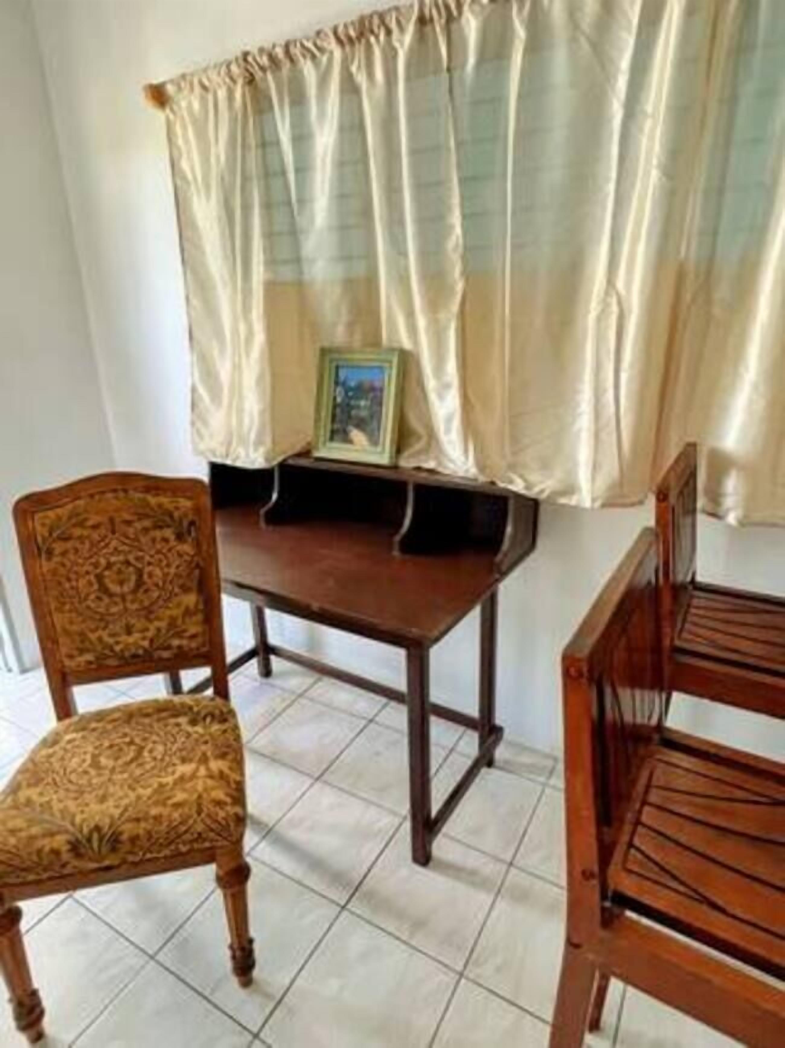 A room with a wooden dining table, two chairs, a small cabinet, and a window with sheer curtains.